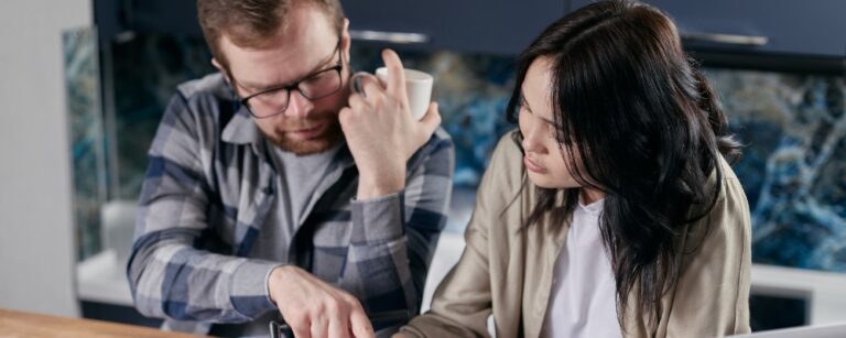 A man and a woman sit at a table in a kitchen, looking at documents together; the man holds a mug and points at the papers.