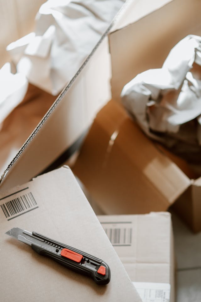 A utility knife rests on a closed cardboard box, surrounded by open boxes and crumpled packing paper.