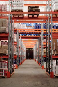 Wide warehouse aisle with tall metal shelving units stocked with various boxes and containers, leading to a closed door at the far end.
