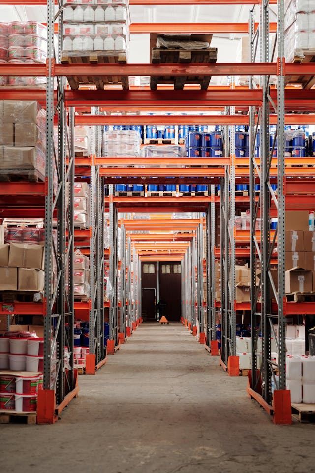 Wide warehouse aisle with tall metal shelving units stocked with various boxes and containers, leading to a closed door at the far end.