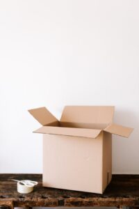 An open cardboard box sits on a wooden table next to a roll of tape and scissors, against a plain white background.