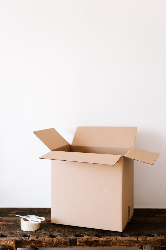 An open cardboard box sits on a wooden table next to a roll of tape and scissors, against a plain white background.